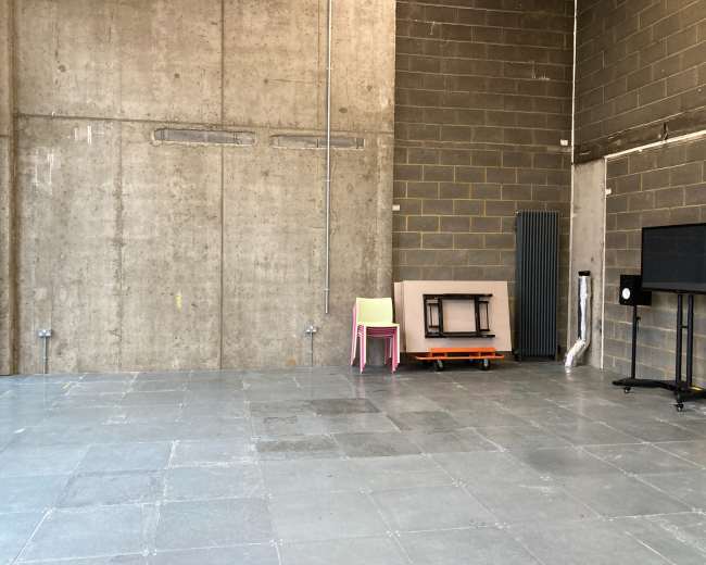 An empty industrial space with a concrete wall, gray tiled floor, and a few pieces of furniture, including stacked chairs and a black television stand.