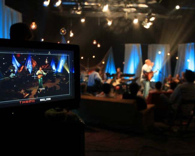 A musician performs on stage while an audience watches, as captured on a video monitor in the foreground.