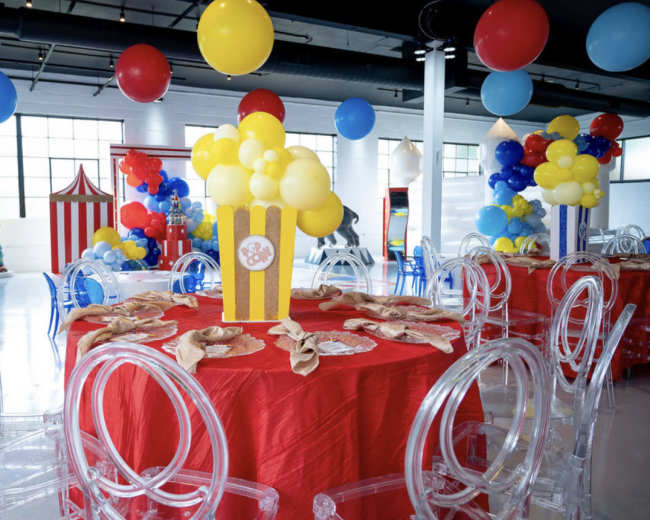 A vibrant party room features circular tables set with yellow and red decor, clear acrylic chairs, and colorful balloon arrangements.