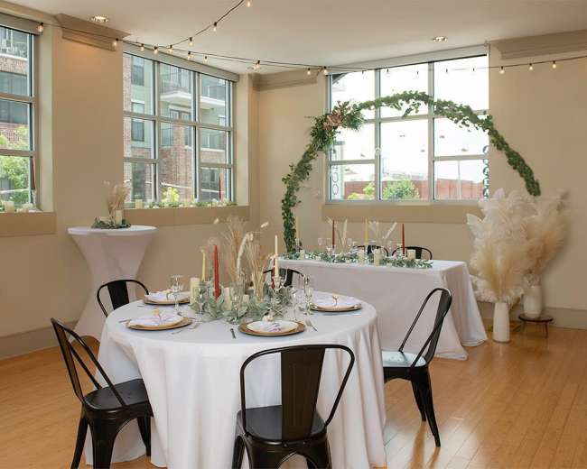A decorated event space with two tables set for dining, featuring candles, floral arrangements, and an arch made of greenery near large windows.