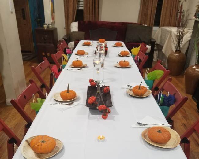 A long table is set with decorative pumpkins on wooden plates, surrounded by red folding chairs and a centerpiece of red decorations.