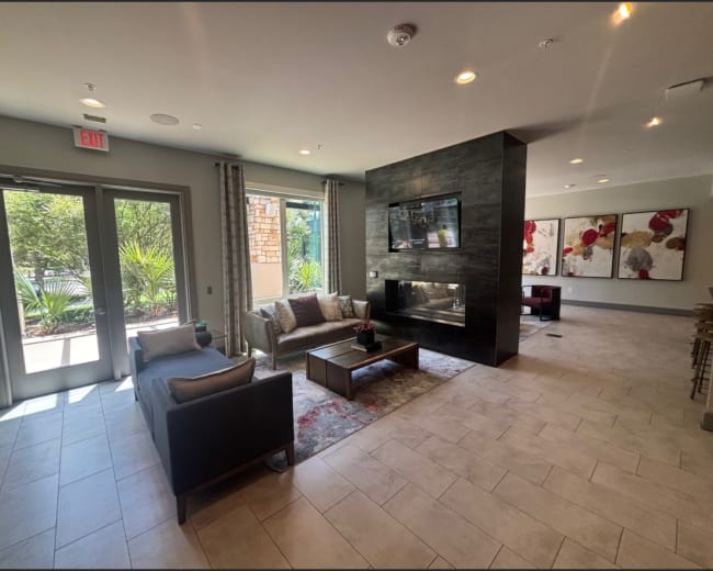 The image shows a modern lounge area with gray tile flooring, a fireplace, a television mounted on a black feature wall, and seating arranged around a small table.