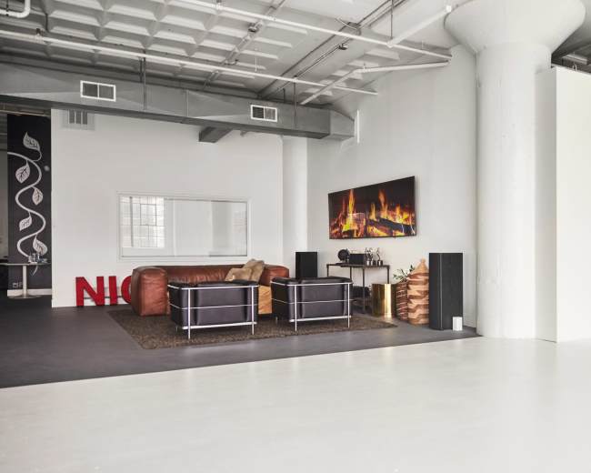 A spacious, modern studio features a seating area with a brown leather couch and two black chairs, accompanied by a large television mounted on the wall displaying a fireplace.