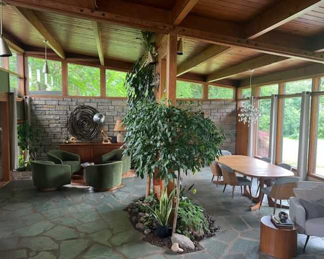 The interior of a spacious, modern living area features large windows, wooden beams, a stone wall, and a mix of green furnishings and plants.