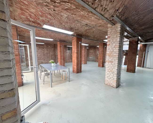 The image shows a large, open industrial space with exposed brick walls, a concrete floor, and a long table with chairs in the center.