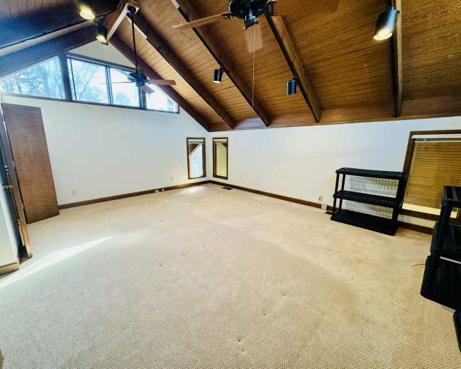 The room features a vaulted wooden ceiling, large windows providing natural light, and a carpeted floor with minimal furniture.
