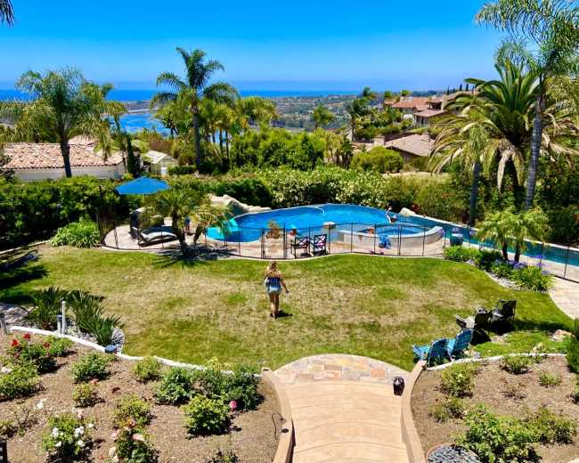 The image shows a backyard with a swimming pool surrounded by palm trees, grassy areas, and a view of the ocean in the background.