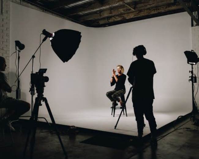 A person sits on a stool in a studio setting, illuminated by multiple lights and surrounded by cameras on tripods.