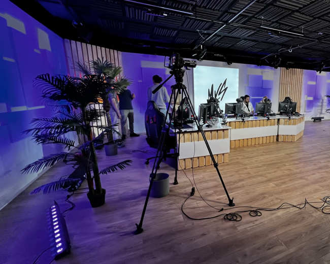 A studio setup features multiple camera tripods, computer stations with monitors, and decorative plants against a backdrop of soft lighting.