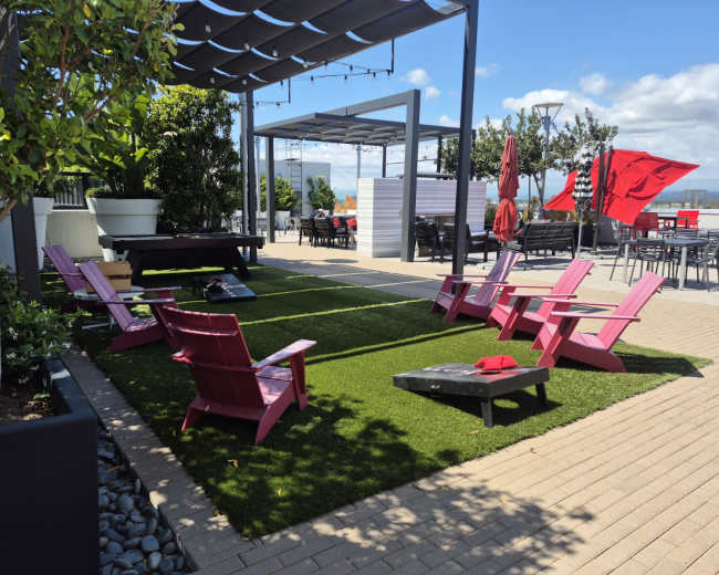 The image shows a landscaped outdoor area with pink lawn chairs arranged on artificial grass, surrounded by potted plants and patio furniture beneath a pergola.