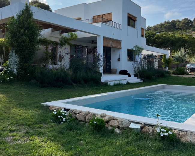 A modern, two-story white villa with a swimming pool and landscaped gardens is set against a hillside.