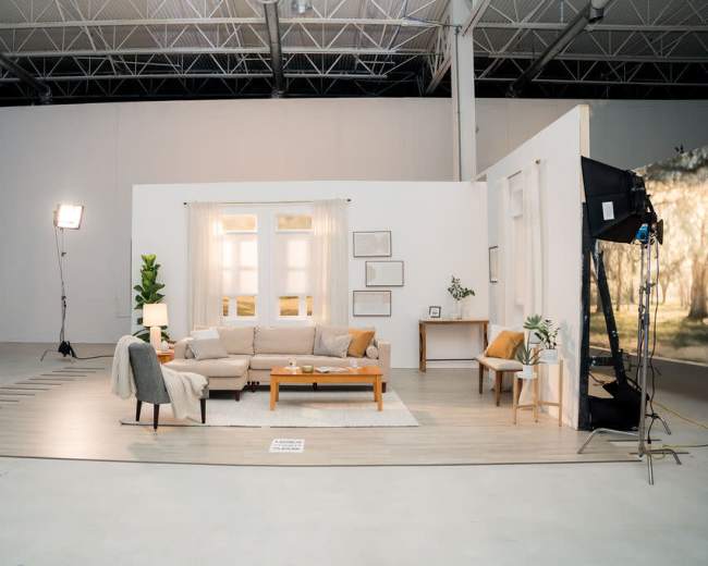 The image shows a staged living room setup with a sofa, coffee table, and decorative plants in a large, well-lit studio space.