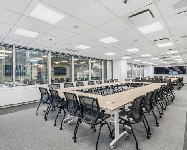 The image shows a modern conference room with a long rectangular table surrounded by black mesh chairs and large windows letting in natural light.