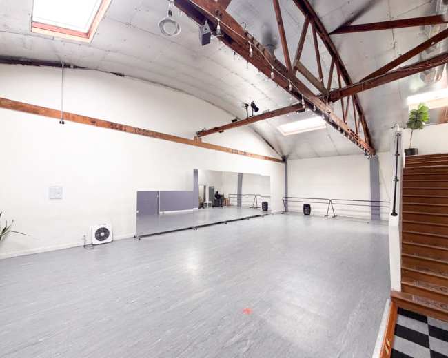 The image shows a spacious studio with a wooden beam ceiling, large mirrors, and wooden stairs leading up to the entrance.