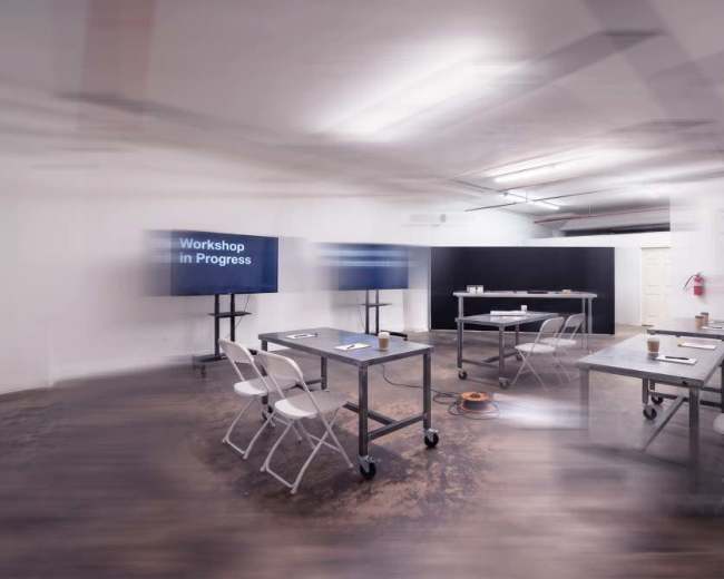 A minimalistic workshop space features several metal tables and chairs arranged around a central area, with two screens displaying "Workshop in Progress" on the walls.