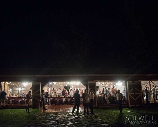 A large gathering takes place inside a barn, illuminated by string lights, with people seated at tables and mingling outside in the dark.