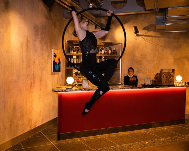 A performer hangs from a hoop suspended from the ceiling, showcasing acrobatic skills in a bar setting with a bartender in the background.