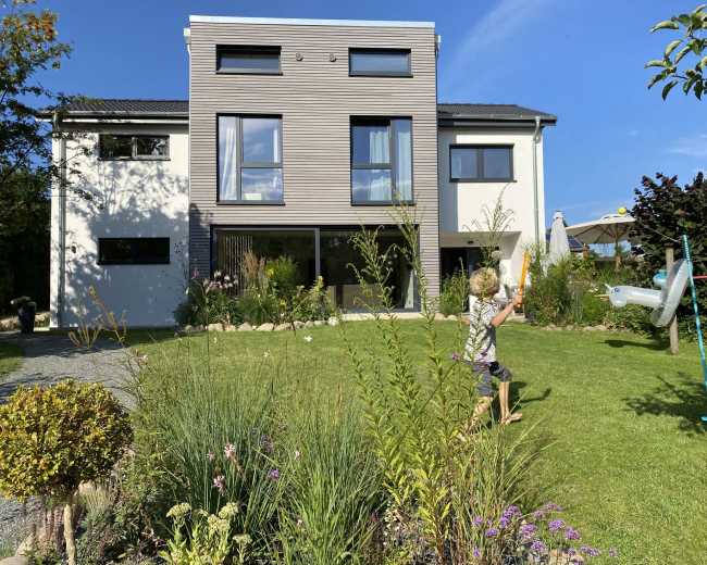A child plays in the garden of a modern two-story house with large windows and a well-maintained lawn.