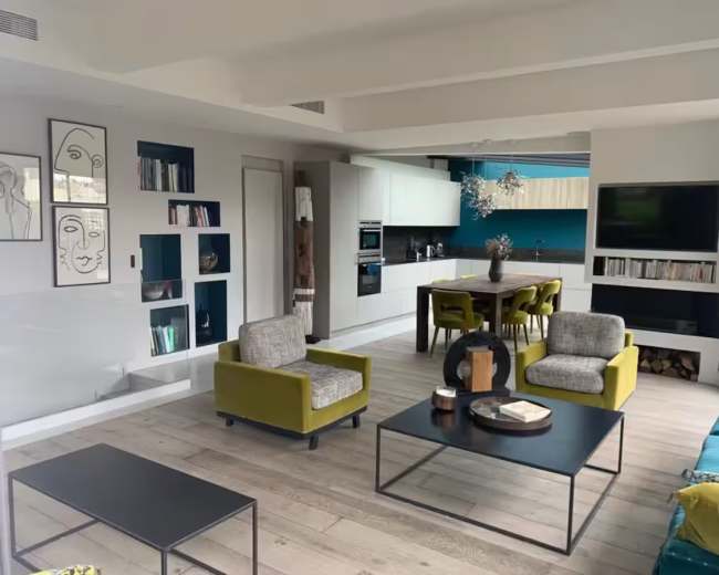 The image shows a modern living space featuring a kitchen area, a dining table, and two armchairs in a light-filled room with wooden flooring and decorative wall art.