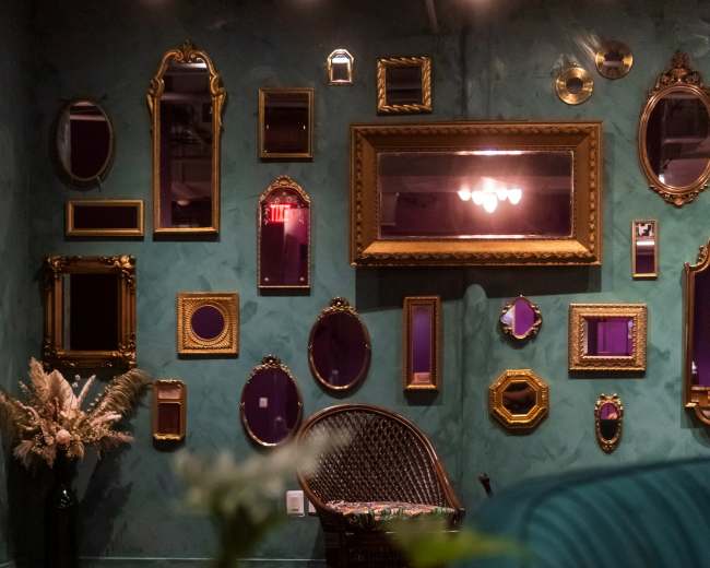 The image shows a room featuring a wall adorned with various ornate mirrors framed in gold, alongside a green couch and a small potted plant in the foreground.