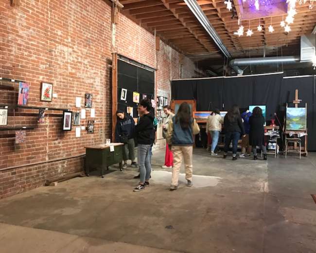 A group of people browse an art exhibition in a spacious, brick-walled venue adorned with various artworks.