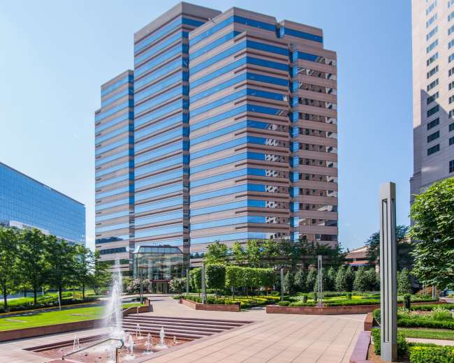 A modern office building with reflective glass windows is surrounded by landscaped gardens and a water fountain.