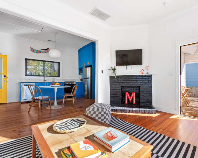 The image shows a modern living space with a kitchen area featuring blue cabinetry, a dining table, a black fireplace with a lit red letter "M," and a door leading to an outdoor deck.
