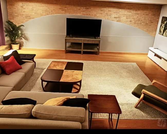 The image shows a spacious living room with a brick wall, a television on a glass cabinet, and a mix of modern furniture including a couch, coffee table, and an armchair.
