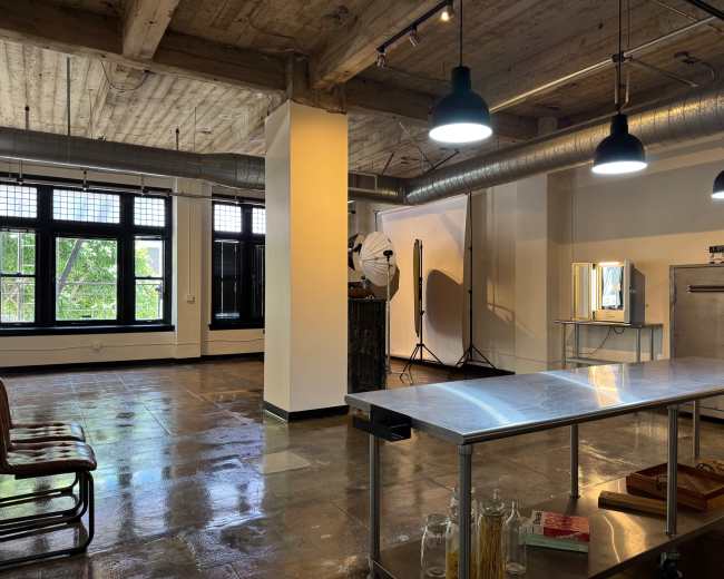 An industrial-style studio features polished concrete floors, large windows, and minimalist furnishings, including a stainless steel table and photography equipment.