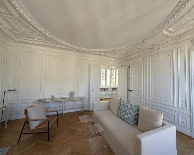 The image shows a bright, elegantly designed living room featuring a white sofa with a decorative pillow, a small coffee table, and ornate wall moldings, complemented by natural light coming through large windows.