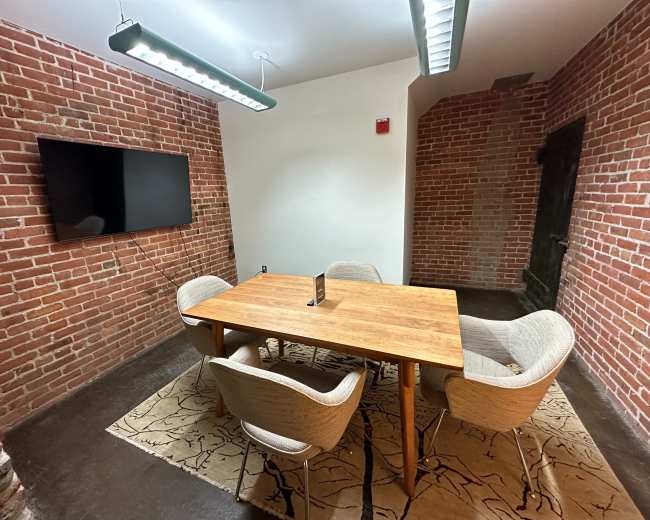 A small meeting room features a wooden table surrounded by four upholstered chairs, a wall-mounted television, and exposed brick walls.