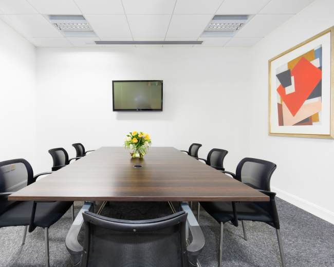 A large conference room features a rectangular table with eight chairs, a wall-mounted TV, and a floral centerpiece.