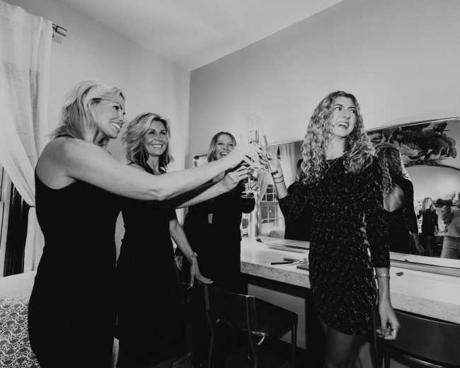 Four women are toasting with champagne glasses in a stylish room, with a mirror reflecting their celebration.