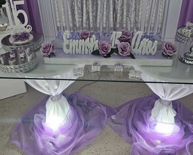 A decorated party area featuring a white throne chair, a purple and silver backdrop, a table setup with decorative items, and a balloon arch.