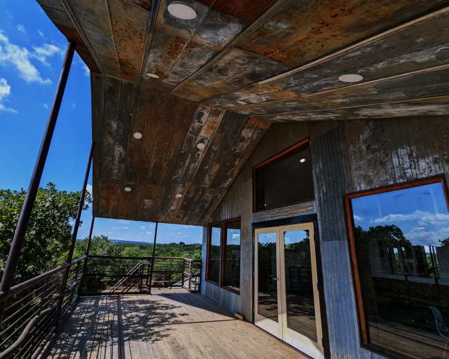 The image displays a modern building with a metal roof and wooden deck overlooking a green landscape under a blue sky.