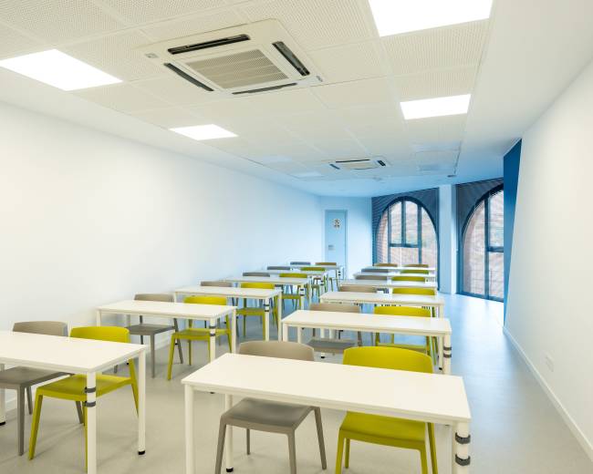 The image shows a modern classroom containing rows of tables and chairs arranged neatly on a light-colored floor.