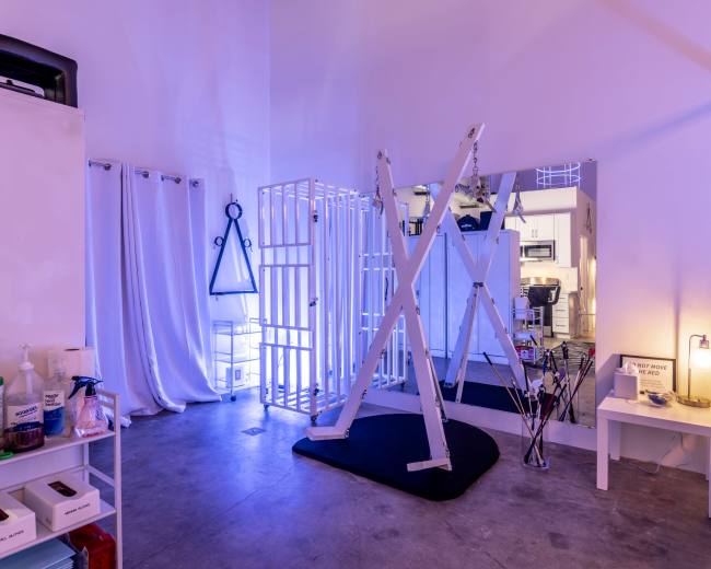 The image shows an indoor space with a minimalist design featuring a large white structure resembling a grid, a mirror, and various objects such as cleaning supplies and a small table.