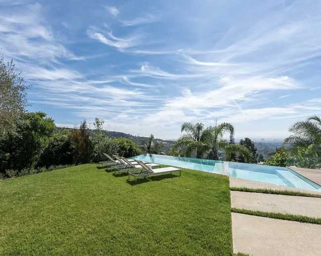 The image shows a modern swimming pool surrounded by green grass, lounge chairs, and palm trees, with a clear blue sky above.