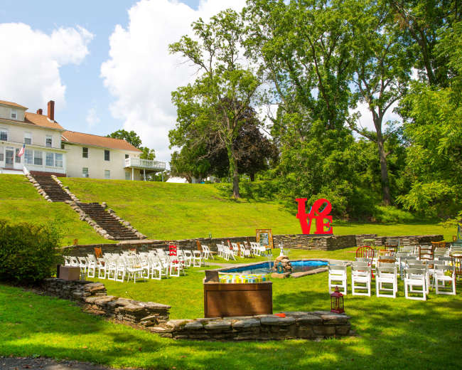 A well-maintained outdoor venue features a large green lawn with white chairs arranged in a semi-circle around a small pool and a large red "LOVE" sign in front of a multi-story white house.