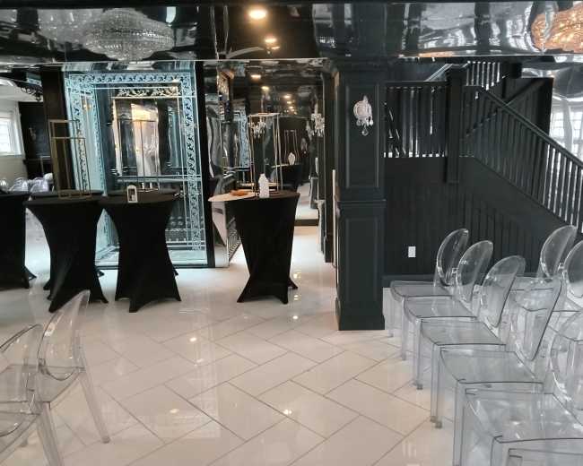 The image shows a stylishly decorated indoor space featuring black and white decor, with tall black tables and transparent chairs arranged for an event, along with a staircase and mirrors on the walls.