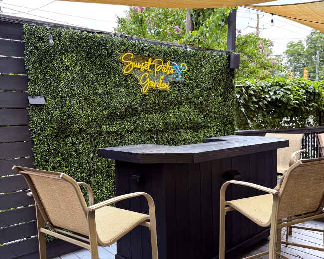 A black outdoor bar is set against a green wall covered in artificial foliage, with the sign "Sunset Patio Garden" illuminated above it.