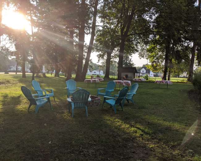 A cluster of blue Adirondack chairs surrounds a fire pit in a grassy area with tables set up in the background.