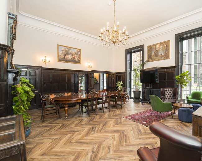The image shows a spacious room with high ceilings, adorned with elegant furniture, plants, and a large chandelier, featuring a dining area and a cozy seating arrangement.