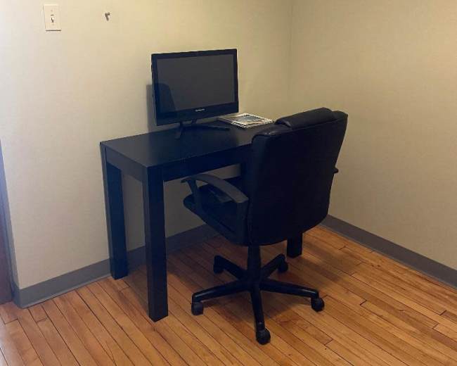 A small corner office setup features a black desk with a computer monitor and a black rolling chair on hardwood flooring.