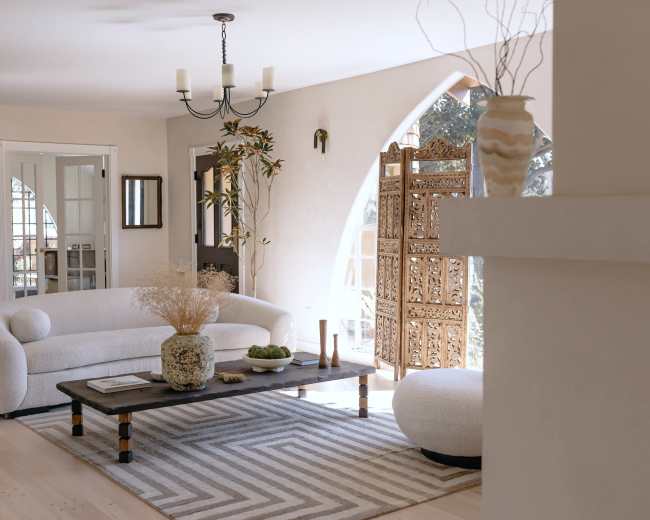 The image shows a bright living room with a curved white sofa, a wooden coffee table with decorative items, and an ornate wooden screen near a large window.