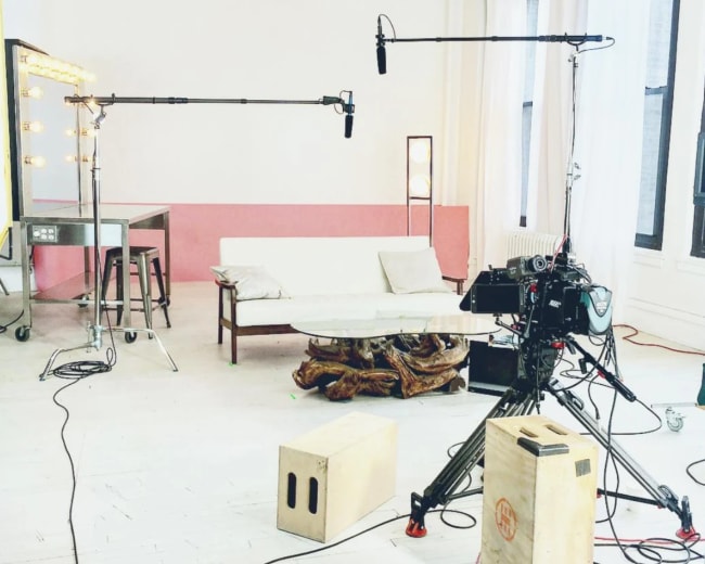 The image shows a staged filming setup with a camera, microphones on stands, a sofa, and lighting equipment in a brightly lit room.