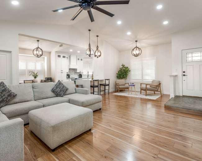 The image shows a modern living room featuring a large sectional sofa, wooden flooring, and stylish light fixtures, with a view into a kitchen area.