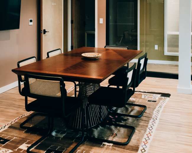 A modern conference room features a wooden table surrounded by black chairs, with a chandelier above and a glass-walled meeting space in the background.