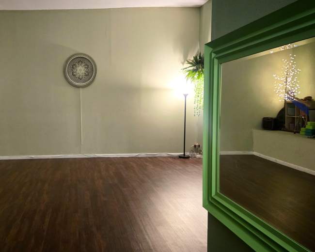 The image shows a minimalist, empty room with a wooden floor, green walls, and a decorative circular wall piece.