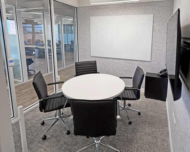 The image shows a small conference room with a round table surrounded by four black chairs, a whiteboard on the wall, and a television mounted on the opposite wall.